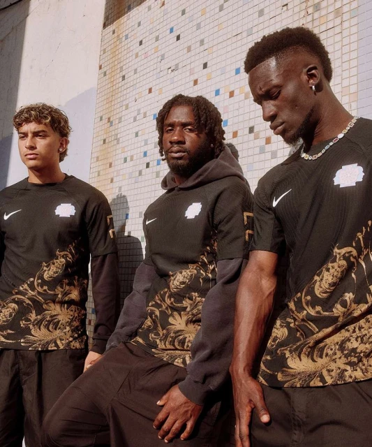 Three male models in athletic streetwear pose against a tiled wall in an urban setting, showcasing black and gold patterned jerseys.