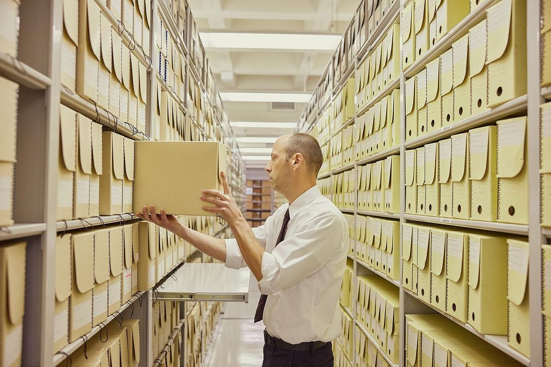 Adult male in a white shirt placing a yellow box on a shelf in a room filled with rows of yellow boxes on metal shelves.
