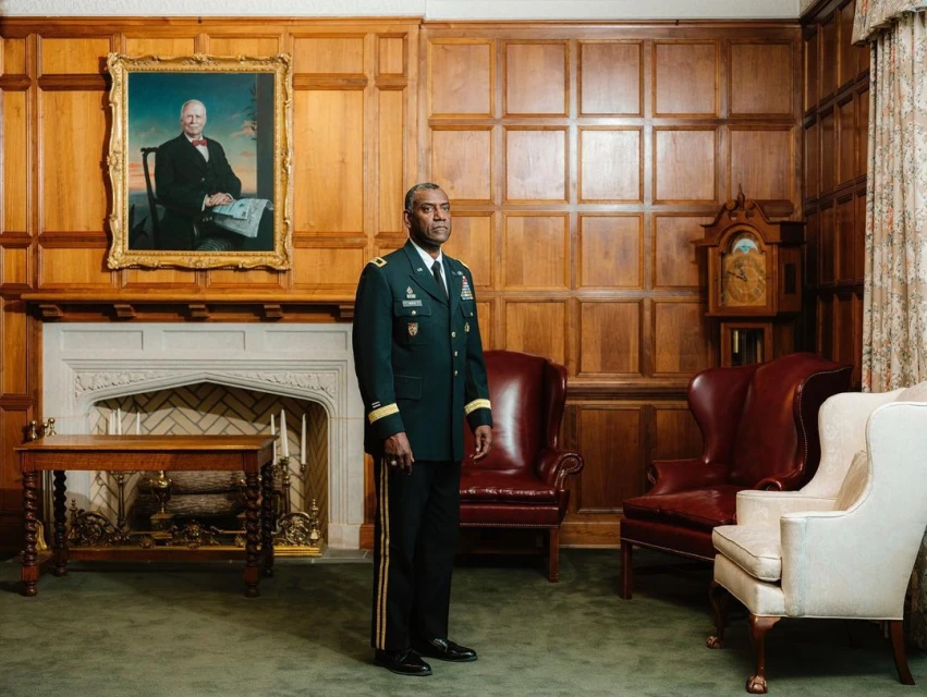 Man in military uniform stands in an ornate room with wood paneling, antique furniture, a fireplace, and a framed portrait.
