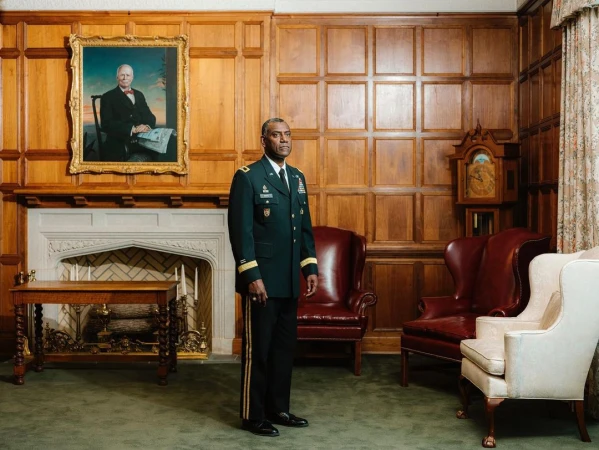 Man in military uniform stands in an ornate room with wood paneling, antique furniture, a fireplace, and a framed portrait.