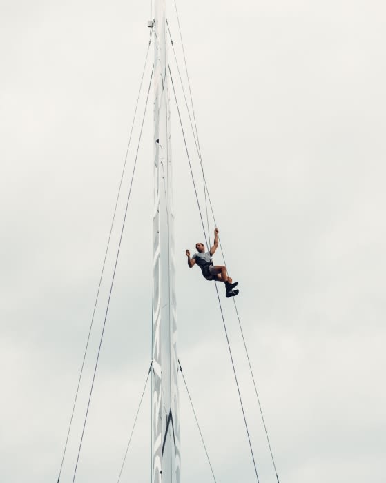 Adult man climbs a tall sailboat mast with rigging. He is wearing shorts and a gray shirt, with a cloudy sky in the background.