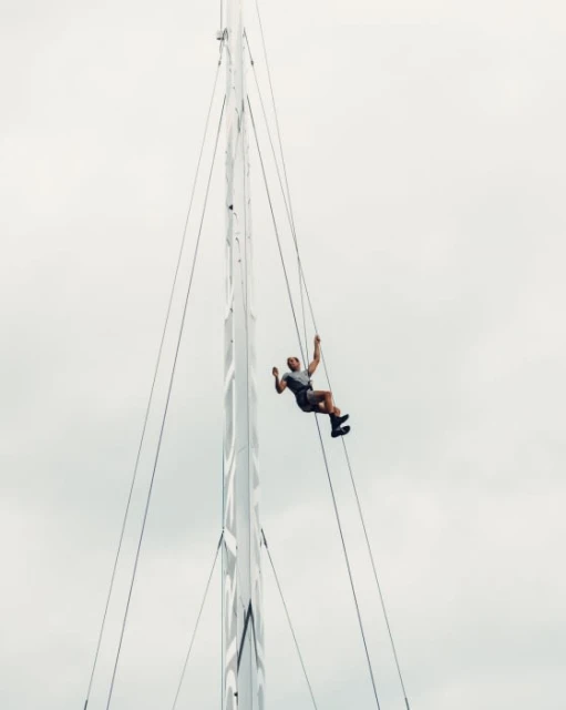 Adult man climbs a tall sailboat mast with rigging. He is wearing shorts and a gray shirt, with a cloudy sky in the background.