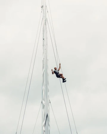 Adult man climbs a tall sailboat mast with rigging. He is wearing shorts and a gray shirt, with a cloudy sky in the background.