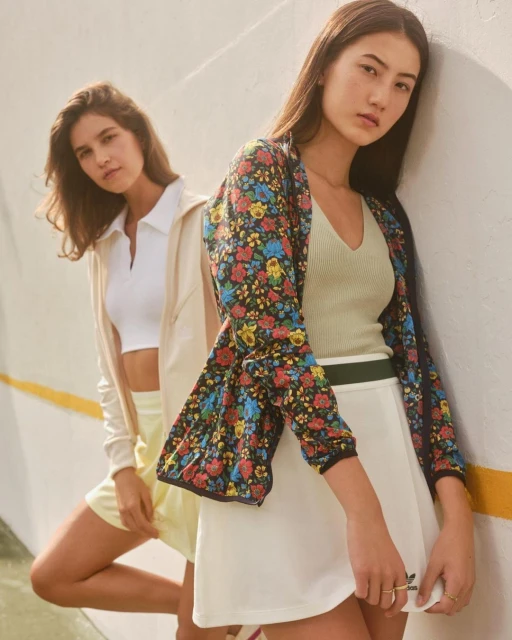 Two women in floral jacket and tennis skirt pose against an off-white wall with yellow stripe; a casual fashion shot.