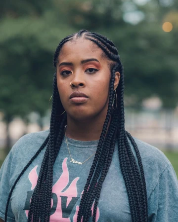 A young African-American woman with braided hair and a nose ring poses outdoors against a blurred green foliage backdrop in medium shot.