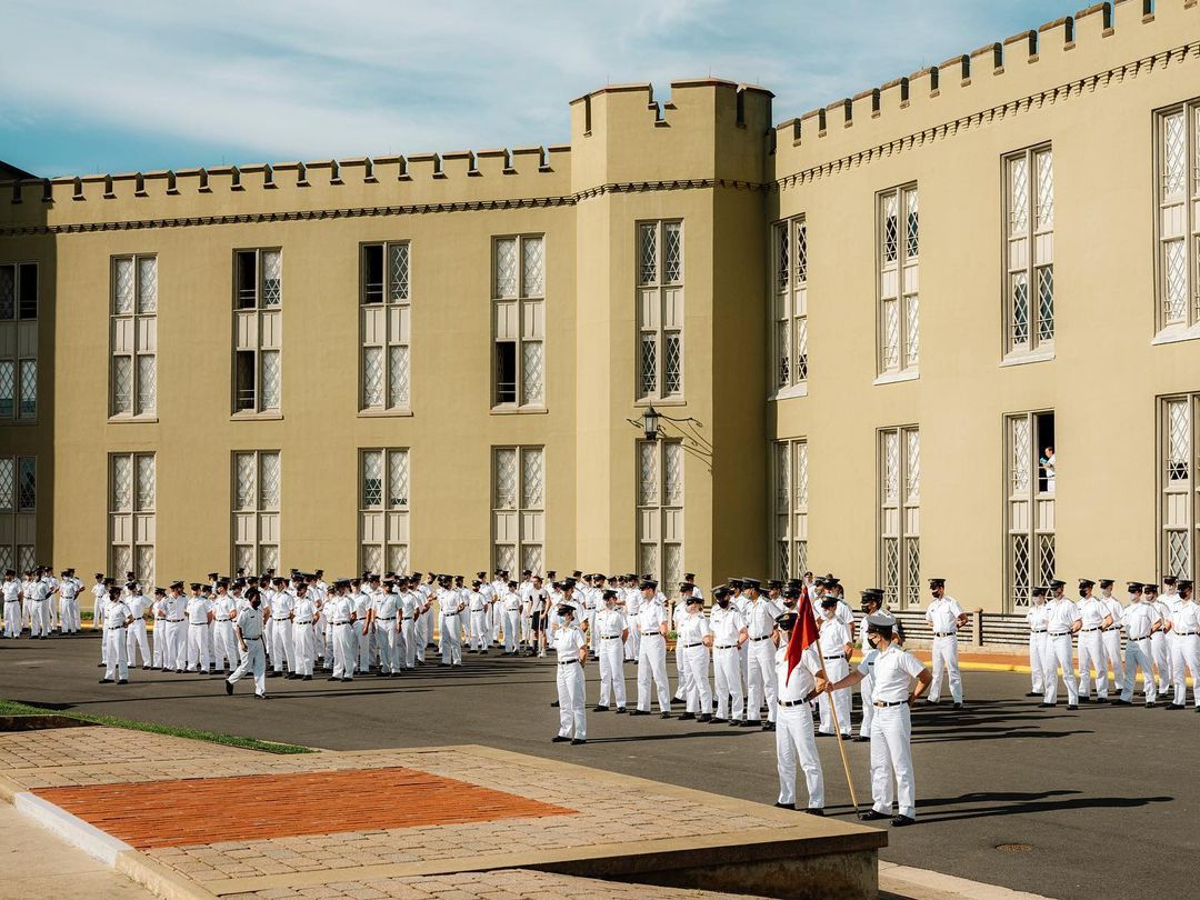 Large group of adult cadets in white uniforms stand at attention in courtyard, classic collegiate architecture and clear blue sky in background.
