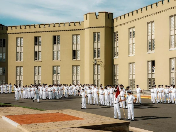 Large group of adult cadets in white uniforms stand at attention in courtyard, classic collegiate architecture and clear blue sky in background.