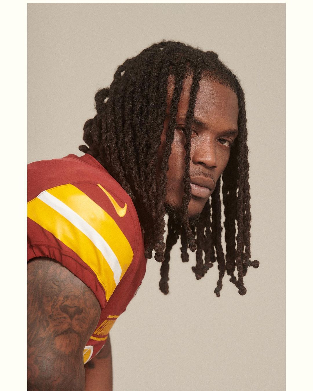 Portrait of an adult male wearing a sports jersey, showcasing his dreadlocks hairstyle, shoulder tattoo, against a neutral backdrop.