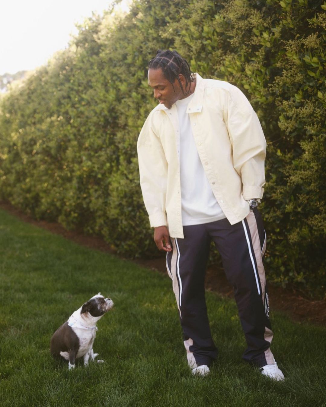 Adult male in a jacket with a dog on the green lawn, posing outdoors with green bushes behind them on a sunny day.