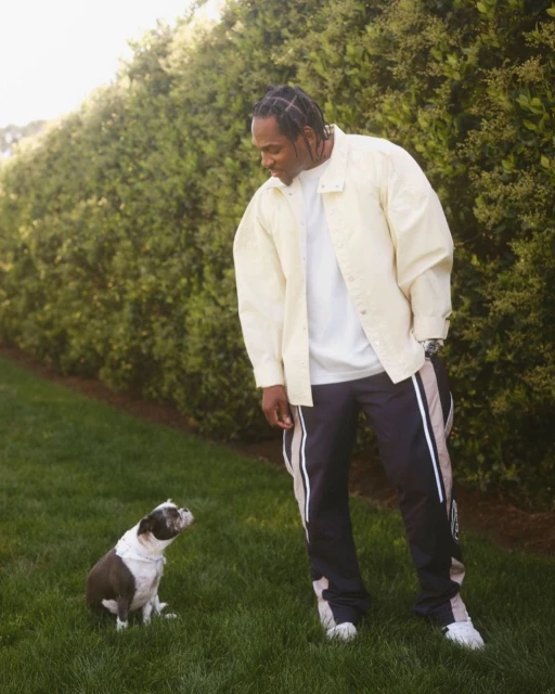 Adult male in a jacket with a dog on the green lawn, posing outdoors with green bushes behind them on a sunny day.