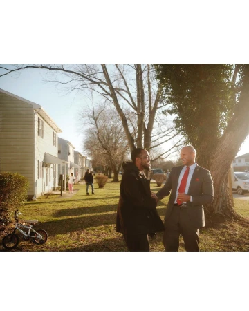 Two adult males shake hands outdoors on a sunny day in front of homes and trees, with a children's bicycle in the foreground.