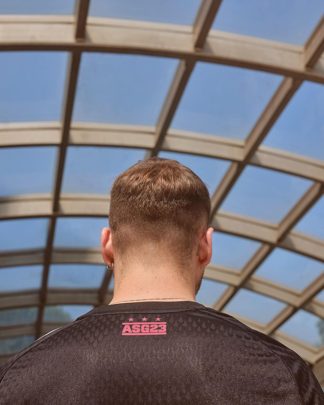 Back of a man's head and shoulders wearing a black sport shirt with pink logo under a skylight providing natural daylight.
