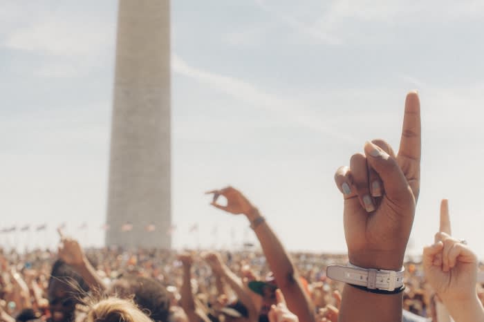 Large crowd with raised hands and fingers pointing up, with Washington Monument in the background against a bright blue sky.