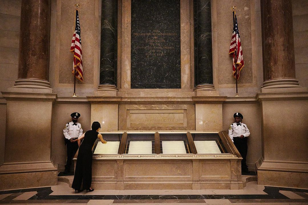 Woman views historical documents in the National Archives museum in Washington, D.C., flanked by American flags and security guards.