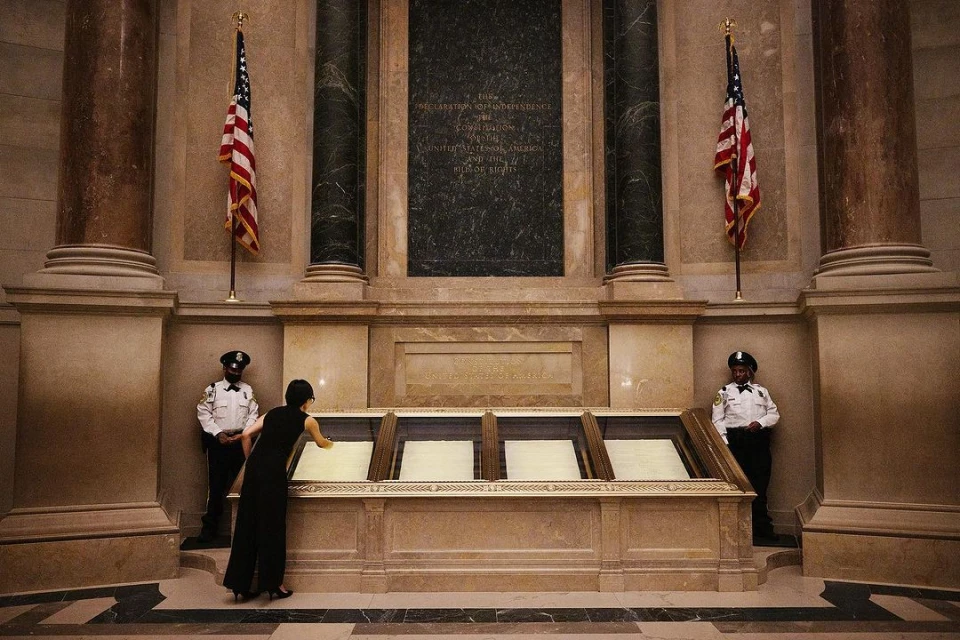 Woman views historical documents in the National Archives museum in Washington, D.C., flanked by American flags and security guards.