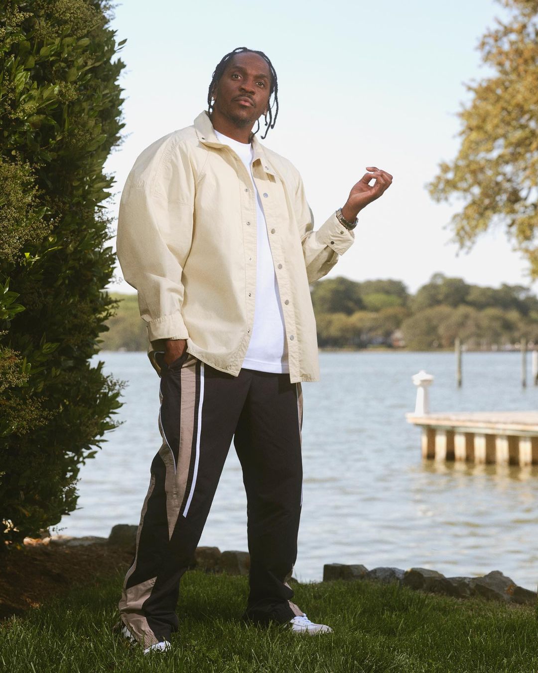Pusha T poses near a waterfront in a beige button-down shirt, white t-shirt, and black pants with beige stripes, exuding casual style.