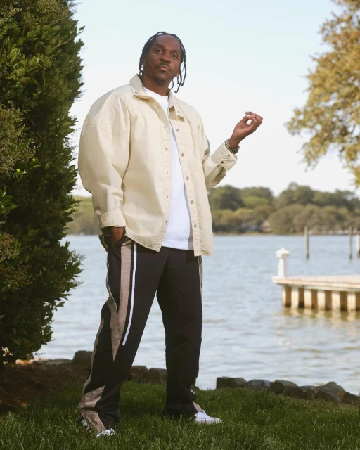 Pusha T poses near a waterfront in a beige button-down shirt, white t-shirt, and black pants with beige stripes, exuding casual style.