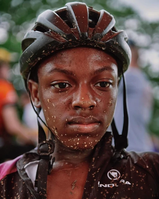 Close-up portrait of a young Black woman cyclist with a helmet, face covered in dirt after a race, set against a blurry outdoor background.
