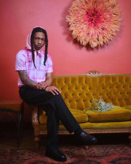 Adult male with braided hair sits on a yellow velvet couch in a photo studio with a pink wall, wearing a pink tie-dye shirt and black pants.