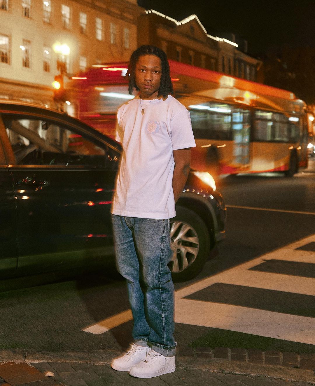 Full-length portrait of a young adult man with dreadlocks, posing on a city street at night with a car and bus in the background.