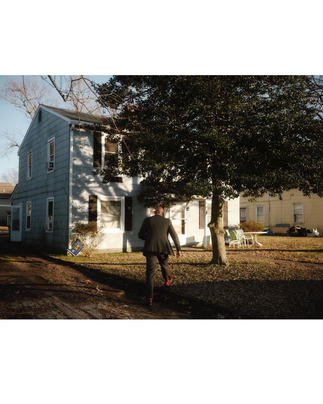Man in a suit walks toward a house under a tree. View from the back showing figure in suit, gray house with dark shutters.