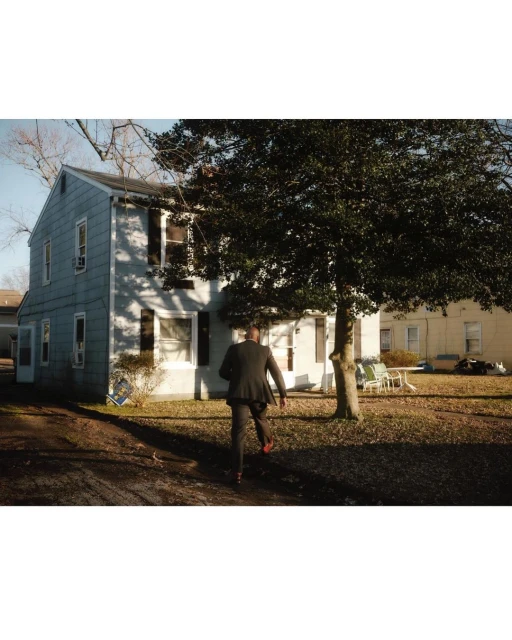 Man in a suit walks toward a house under a tree. View from the back showing figure in suit, gray house with dark shutters.