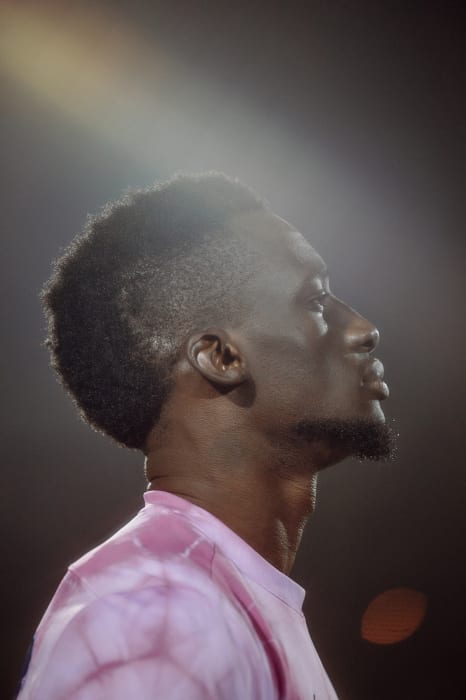 Portrait of a young Black man with a beard looking up towards the light, wearing a tie-dye T-shirt in a dark, shadowed environment.