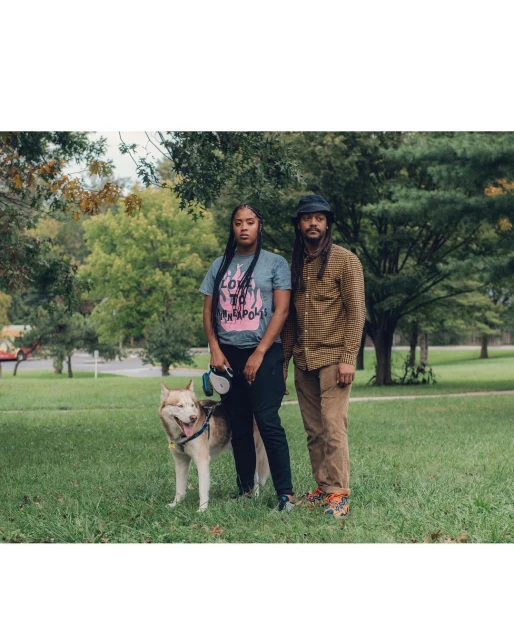 A medium shot of a stylish couple posing with a husky in a grassy urban park with trees and a road visible in the background.