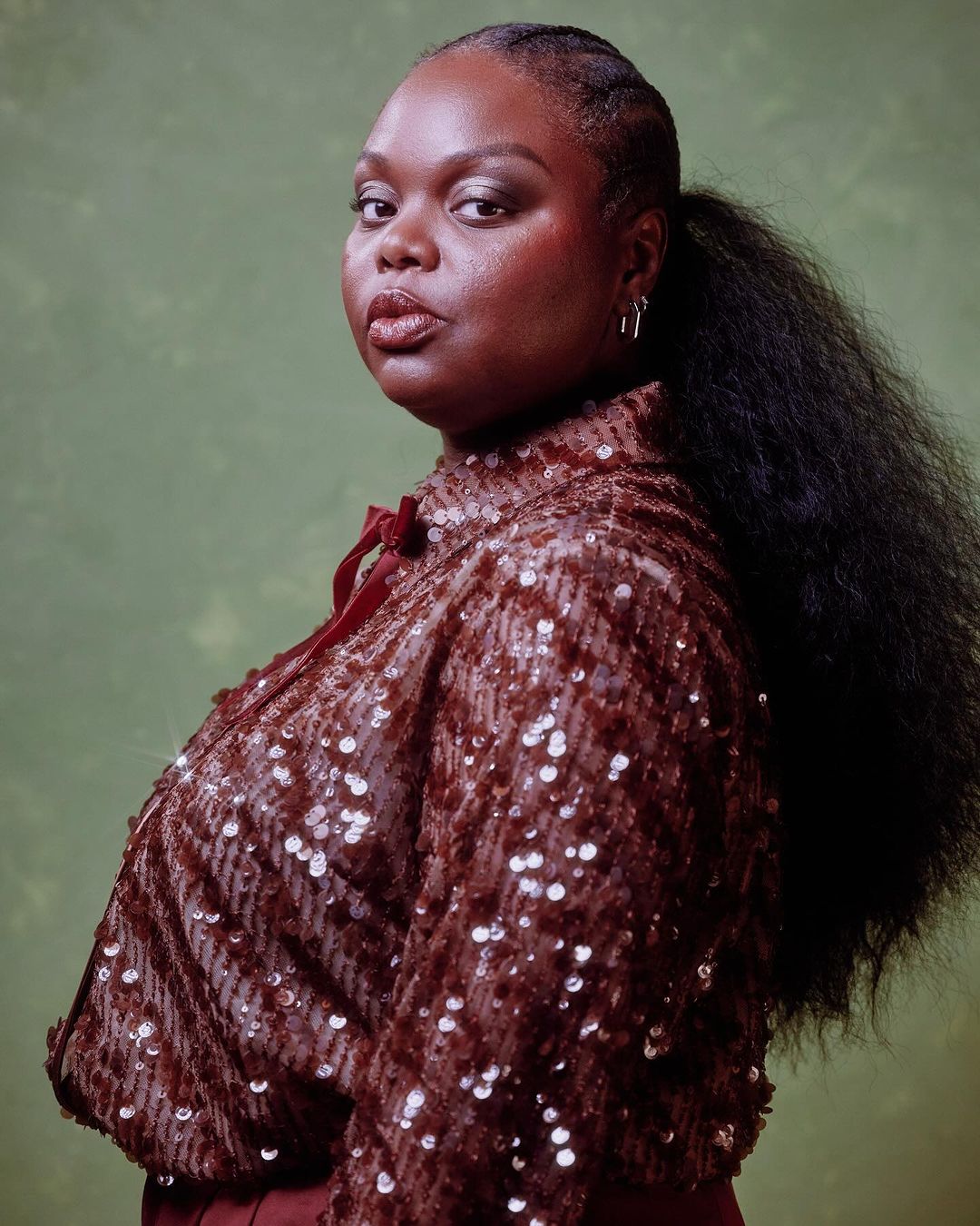 Portrait of a black woman with dark brown sequin top and ponytail against a mottled green background in a studio setting. Beauty and fashion.