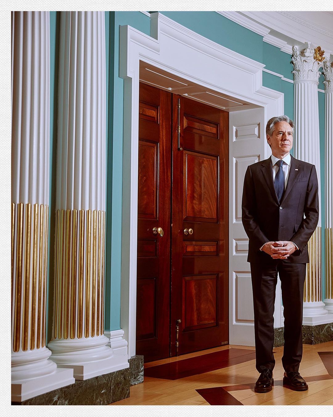 Full-length portrait of a male in a suit standing in front of a set of large, dark wood doors in an ornate, architectural setting.