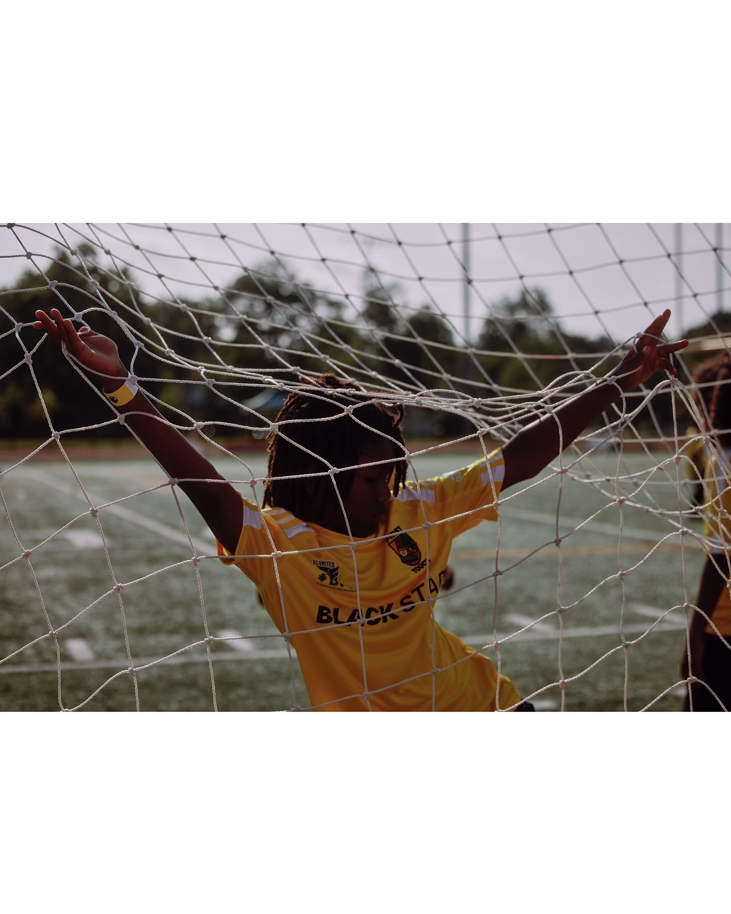Young adult soccer player celebrates in a net, wearing a yellow jersey with 'Black Star' text, on a green field in daylight.