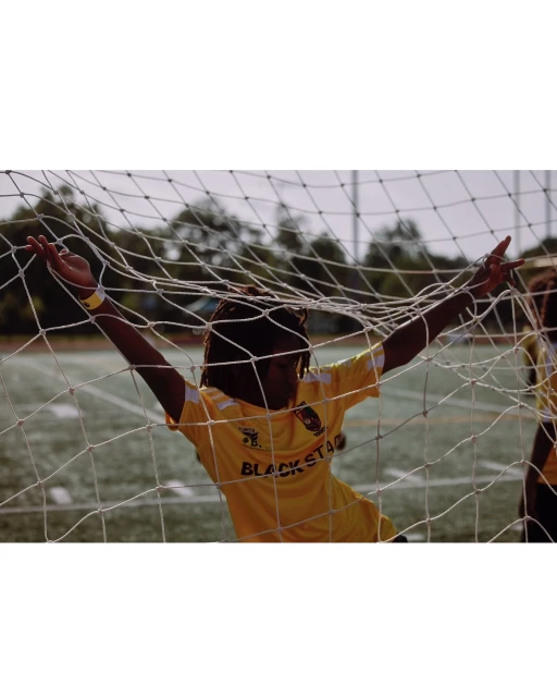 Young adult soccer player celebrates in a net, wearing a yellow jersey with 'Black Star' text, on a green field in daylight.