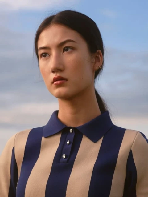 Portrait of a young adult woman looking away from the camera, wearing a blue-and-beige striped polo shirt against a sky background.