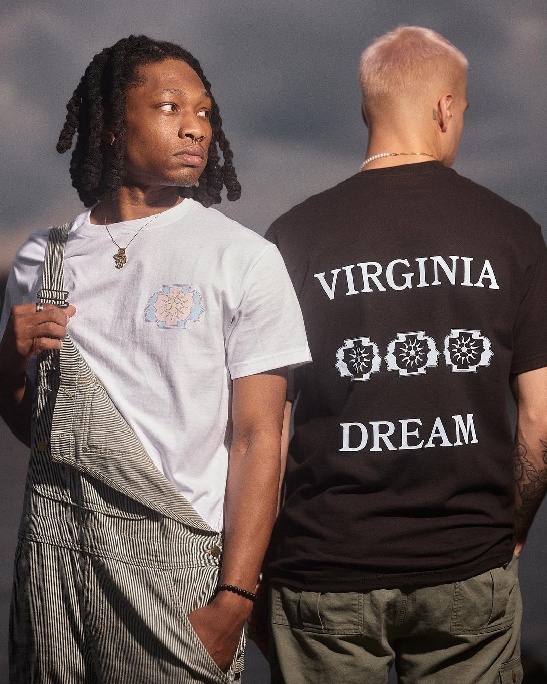 Two people wearing branded t-shirts pose outdoors against an overcast sky, showcasing casual fashion and streetwear style in natural light.