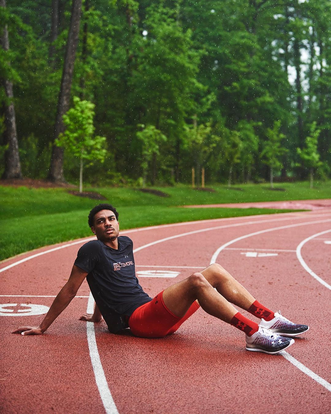 Male athlete rests on a red running track outdoors in daytime, wearing running shoes, red shorts and socks, and a dark T-shirt.