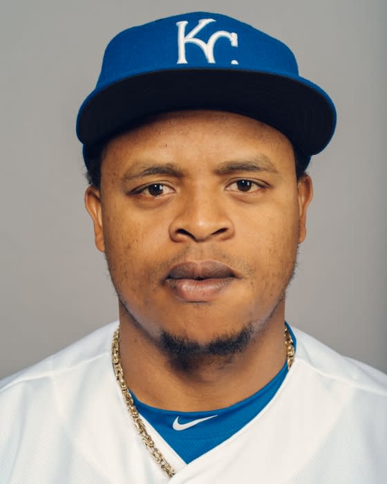 Close-up shot of an adult male wearing a Kansas City Royals baseball cap and gold chain against a grey background.