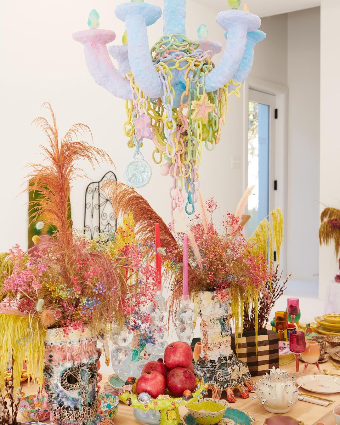 Pastel chandelier hangs over a colorful tabletop arrangement with decorative vases, pomegranates, and other artistic elements indoors.