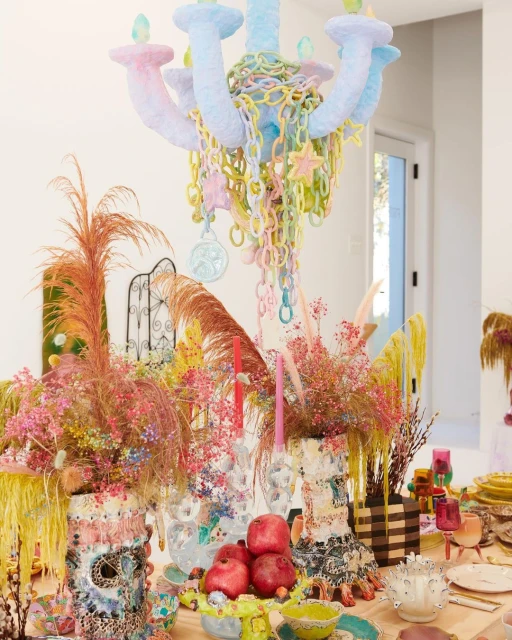 Pastel chandelier hangs over a colorful tabletop arrangement with decorative vases, pomegranates, and other artistic elements indoors.