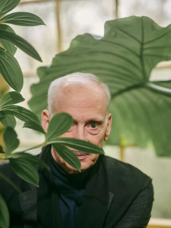 Portrait of an elderly man peering through green leaves in a greenhouse setting, wearing a dark jacket and scarf.
