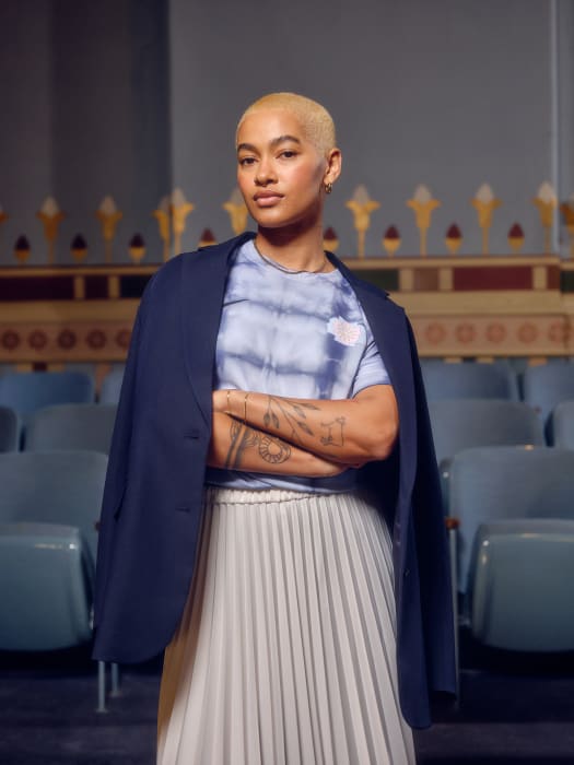 Woman in blazer and pleated skirt poses in theater with arms crossed, exuding modern elegance in the vibrant, light-filled scene.