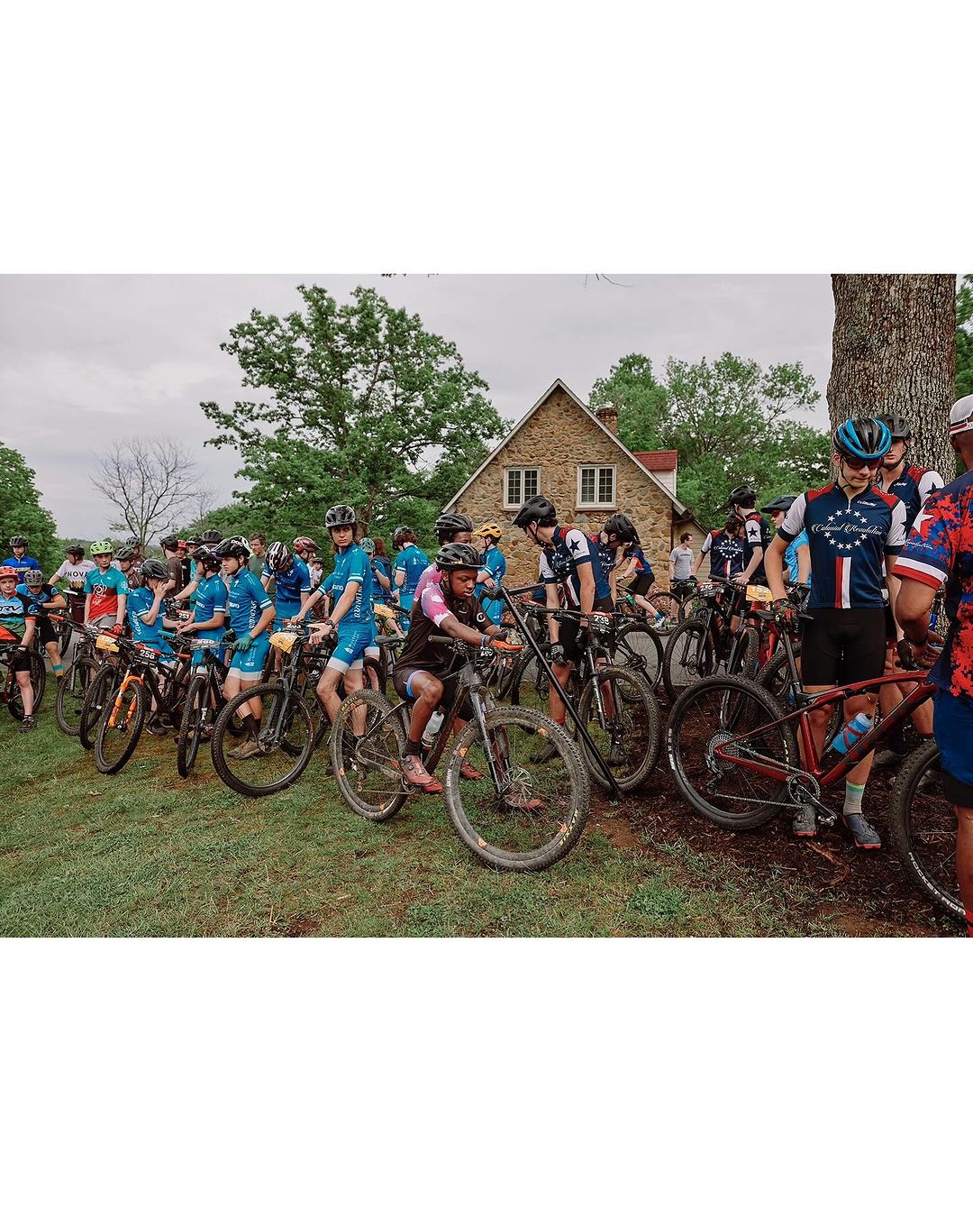 Group of cyclists on mountain bikes gather outdoors near a stone house on a cloudy day, showcasing a cycling event and rural setting.