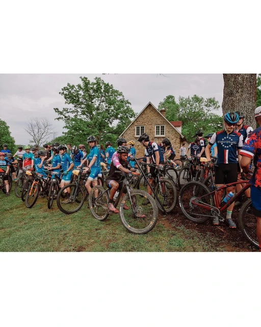Group of cyclists on mountain bikes gather outdoors near a stone house on a cloudy day, showcasing a cycling event and rural setting.