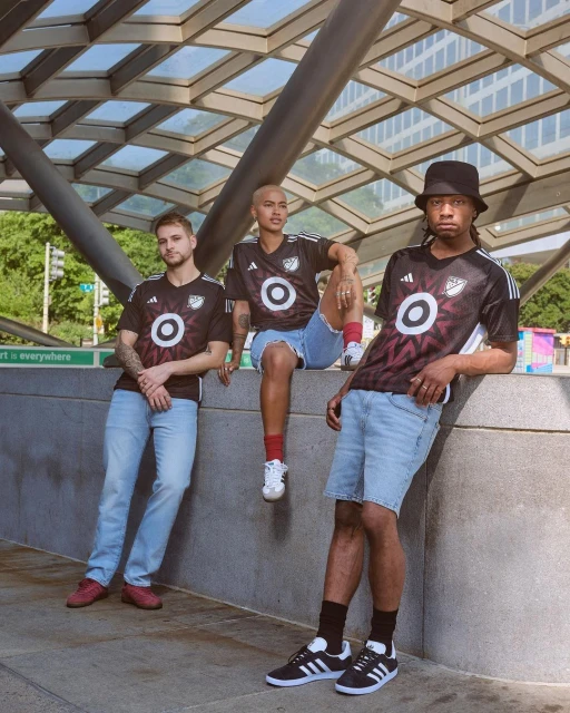 Three adult people in sports jerseys pose under a glass and steel canopy, wearing shorts and sneakers, in an urban outdoor setting. 
