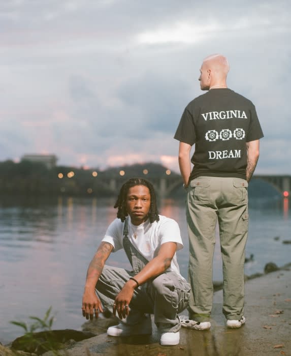 Two people pose near a river. One is crouched on a stone wearing overalls, the other stands in a t-shirt and pants, facing away.