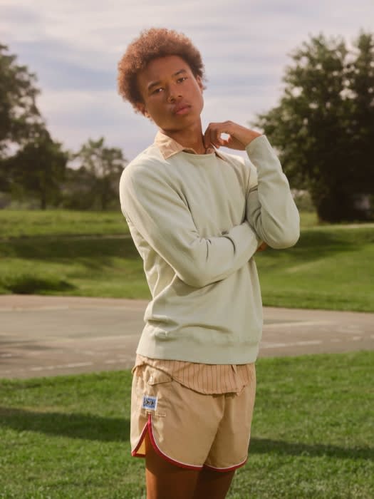 Man with afro wearing light green sweater and khaki shorts poses outside on a sunny day with grassy background and light blue sky.