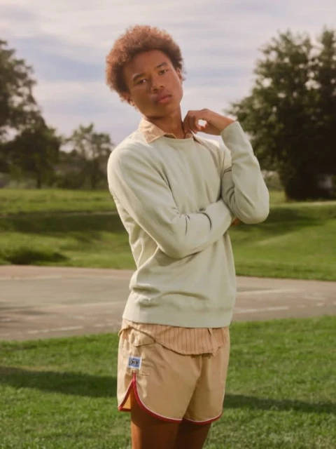 Man with afro wearing light green sweater and khaki shorts poses outside on a sunny day with grassy background and light blue sky.