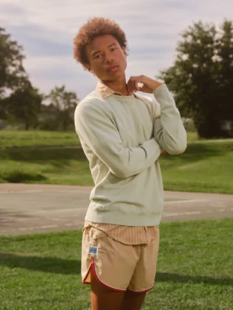 Man with afro wearing light green sweater and khaki shorts poses outside on a sunny day with grassy background and light blue sky.