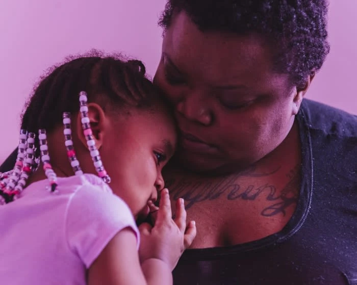 Close-up studio shot of an African-American mother embracing her young daughter, showcasing family love and bonding in soft, pink lighting.