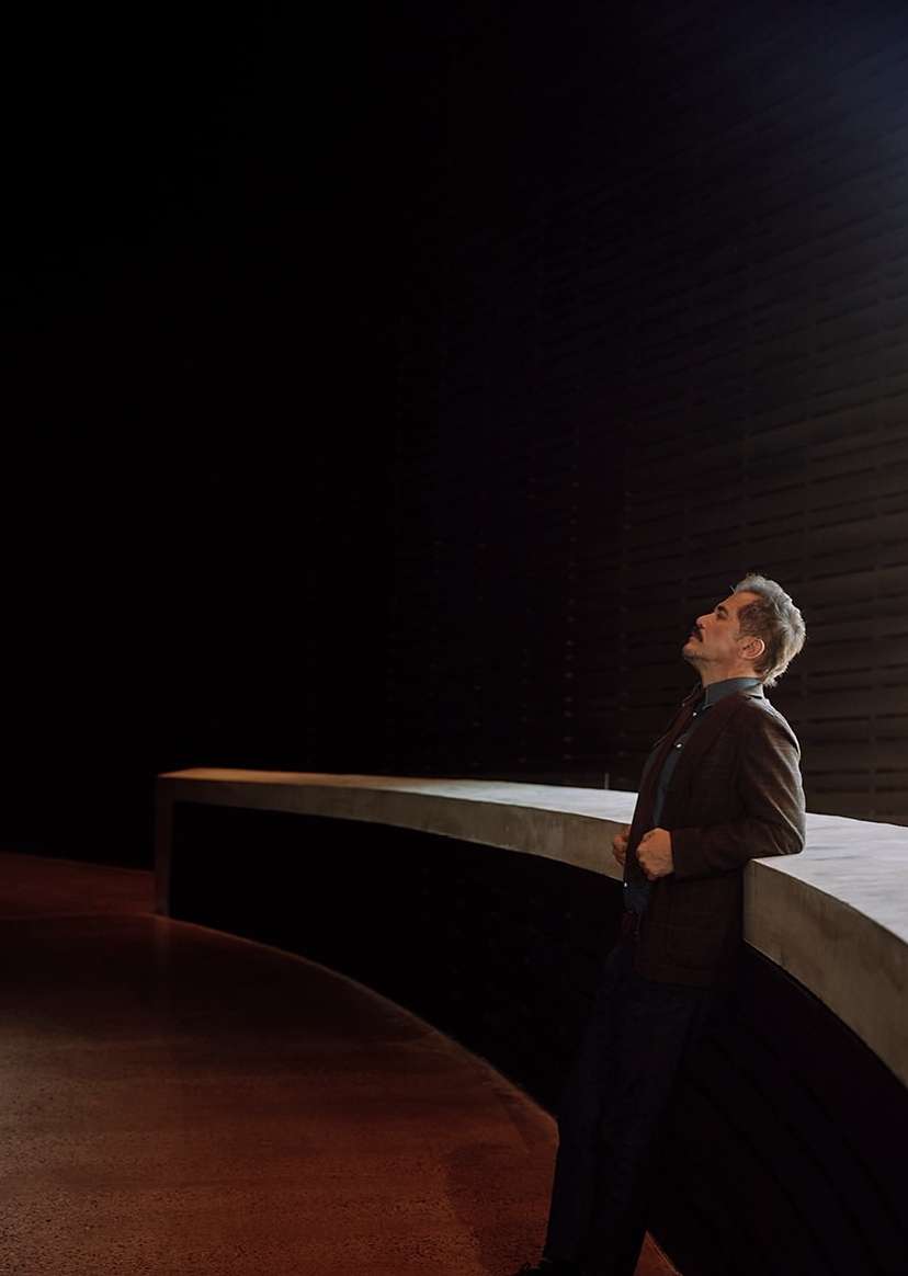 Man in jacket leans on concrete structure in a dimly lit space, looking upward. Interior architecture is minimalist with dark background.