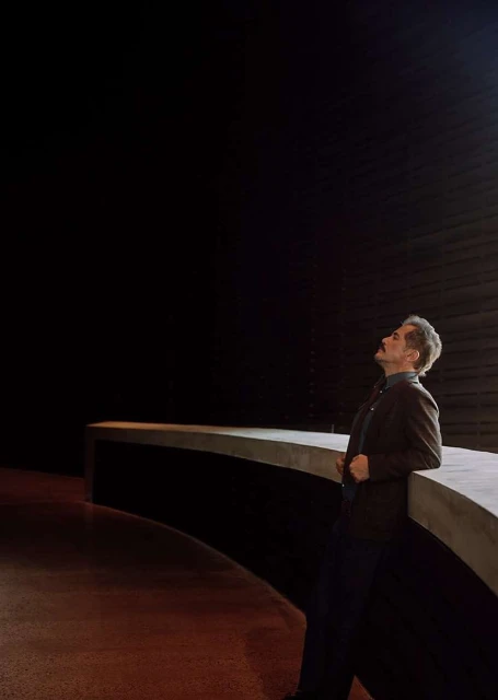 Man in jacket leans on concrete structure in a dimly lit space, looking upward. Interior architecture is minimalist with dark background.